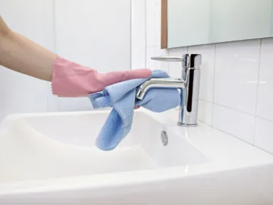 Woman cleaning hard water stain off the faucet with microfiber cloth and gloves