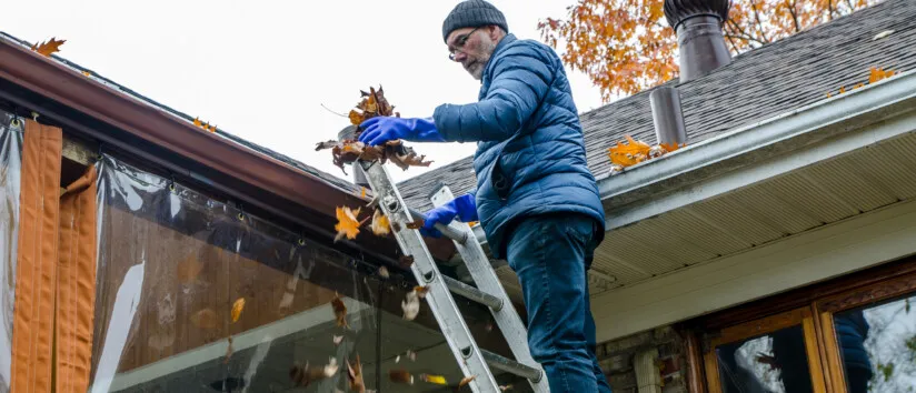 Man on ladder removing leaves from gutter practicing proactive home maintenance