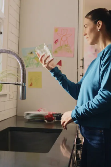 a woman holding a glass of water near the sink looking outside on a sunny day