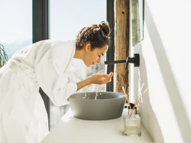 Young woman in white bath cleaning face over sink to minimize hard water affects on hair, skin, and nails