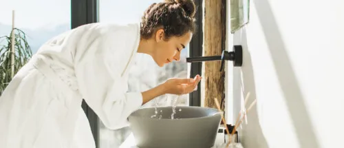 Young woman in white bath cleaning face over sink to minimize hard water affects on hair, skin, and nails